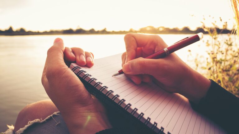 Close up hand of young woman with pen writing on notebook at riv Close up hand of young woman with pen writing on notebook at riverside in the evening.